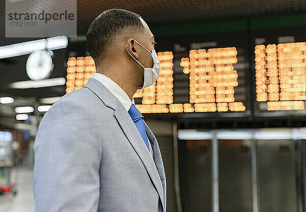 Geschäftsmann mit Schutzmaske steht vor der Abfahrtstafel am Bahnhof