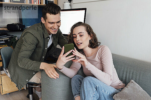 Junge Frau teilt Smartphone mit Freund  der auf einem Stuhl im Wohnzimmer sitzt