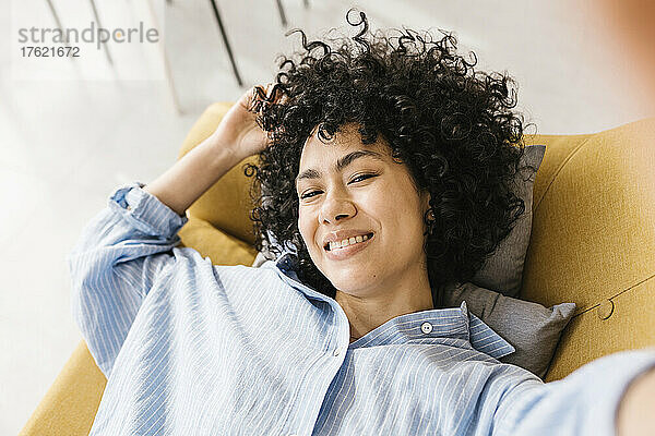 Lächelnde junge Frau mit schwarzen Locken  die zu Hause auf dem Sofa liegt