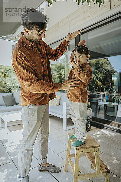 Father measuring height of son standing on stool in patio