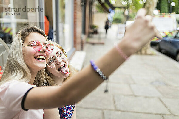 Cheerful teenage girl taking selfie through mobile phone with friend sticking out tongue