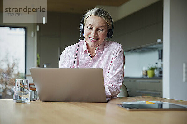 Lächelnde blonde Frau mit Kopfhörern und Laptop am Tisch sitzend