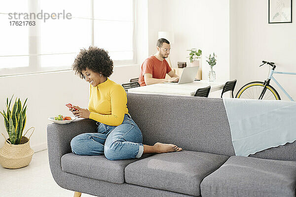 Lächelnde junge Frau benutzt Smartphone  während Mann mit Laptop im Wohnzimmer sitzt