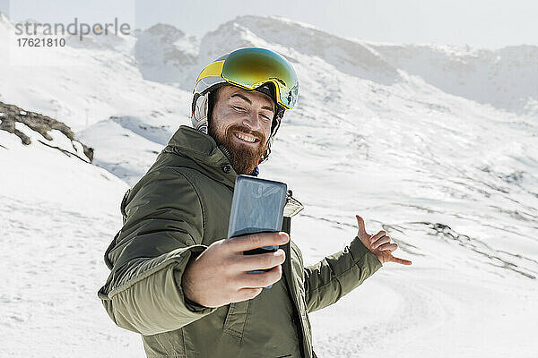 Lächelnder junger Mann gestikuliert und macht an einem sonnigen Tag ein Selfie mit dem Mobiltelefon