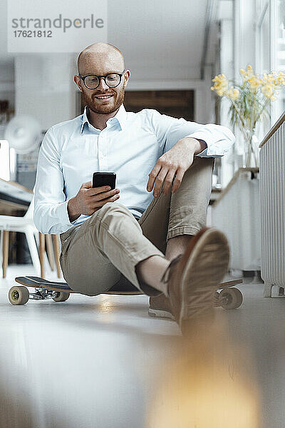 Lächelnder junger Geschäftsmann  der im Büro auf dem Skateboard sitzt und sein Mobiltelefon benutzt