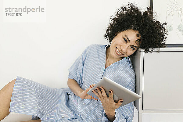 Happy young woman with tablet PC leaning on cabinet at home