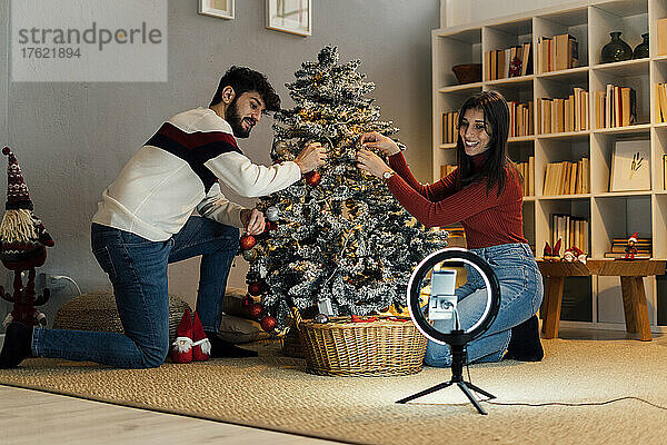 Glückliches Paar schmückt den Weihnachtsbaum und vloggt über sein Mobiltelefon  das zu Hause an einer Ringleuchte befestigt ist