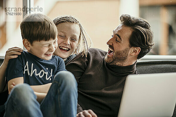 Fröhlicher Vater und Söhne sitzen mit Laptop und lachen zu Hause
