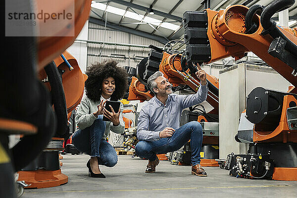 Ingenieur sitzt mit einem Tablet-PC neben dem Entwickler und zeigt auf den Roboterarm in der Fabrik