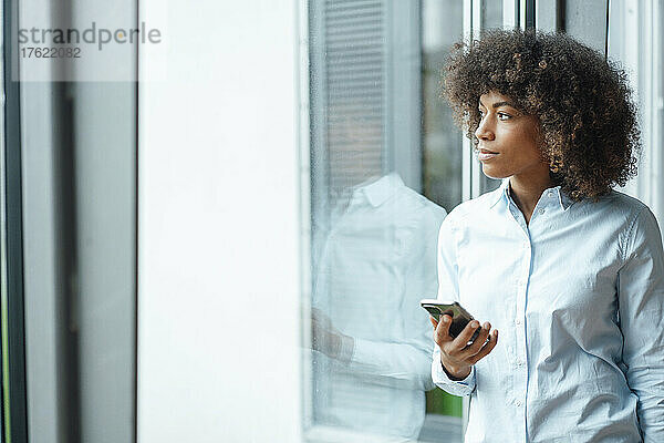 Geschäftsfrau schaut mit Mobiltelefon durchs Fenster im Büro
