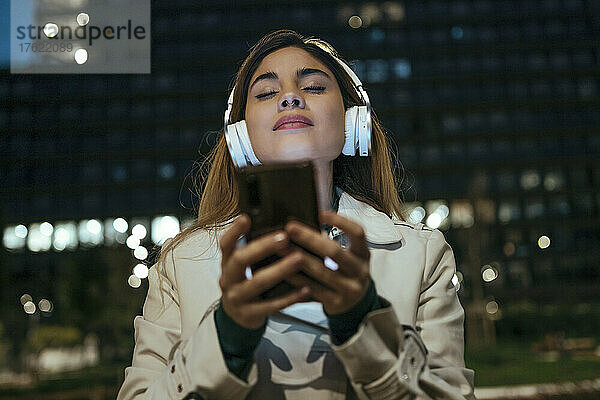 Frau mit geschlossenen Augen hält ihr Mobiltelefon in der Hand und hört nachts Musik über Kopfhörer