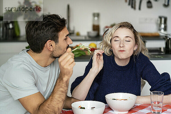 Junger Mann blickt mit geschlossenen Augen auf seine Freundin  die am Tisch isst