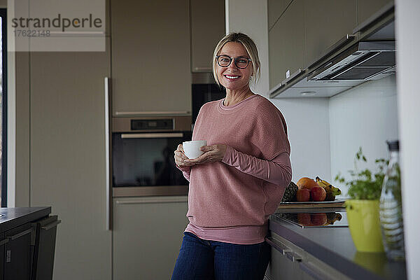 Glückliche blonde Frau hält eine Kaffeetasse in der Hand und steht zu Hause an der Küchentheke