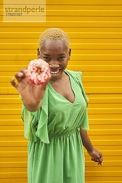 Cheerful young woman holding doughnut in front of yellow wall