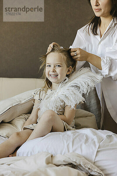 Frau bindet die Haare ihrer fröhlichen Tochter  die zu Hause mit Feder auf dem Bett sitzt