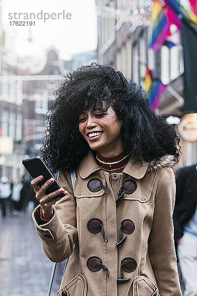 Glückliche junge Frau mit zerzausten Haaren surft per Smartphone im Netz in der Stadt