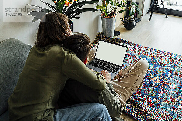 Junge Frau sitzt mit dem Arm um ihren Freund und benutzt Laptop zu Hause