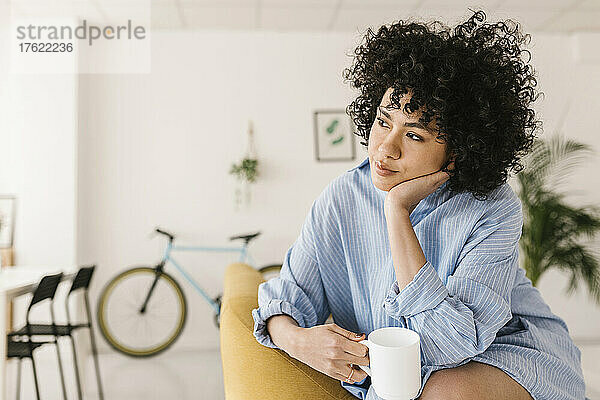 Nachdenkliche junge Frau sitzt mit Kaffeetasse im Wohnzimmer zu Hause