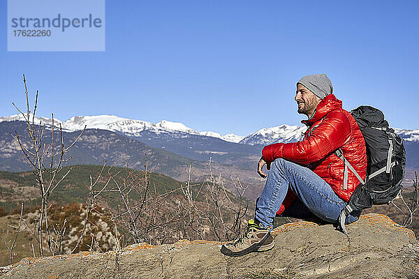 Lächelnder Mann mit Rucksack sitzt auf einem Felsen