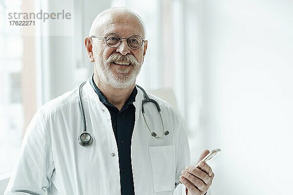 Lächelnder Oberarzt mit Smartphone im Krankenhaus