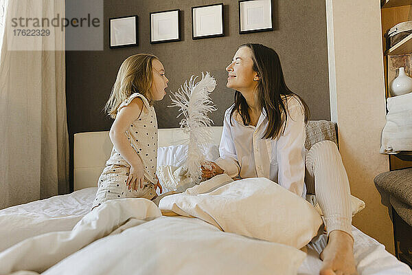 Verspieltes Mädchen bläst auf Federn  gehalten von einer Frau  die zu Hause mit geschlossenen Augen auf dem Bett sitzt