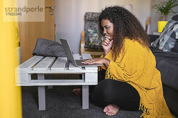 Junge Frau mit lockigem Haar sitzt mit gekreuzten Beinen und benutzt Laptop am Couchtisch im Wohnzimmer