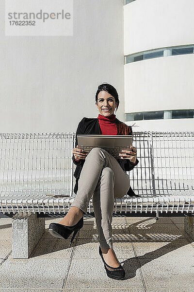 Lächelnde Geschäftsfrau mit Laptop sitzt auf Bank