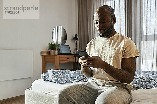 Mann benutzt Smartphone und sitzt zu Hause im Schlafzimmer auf dem Bett