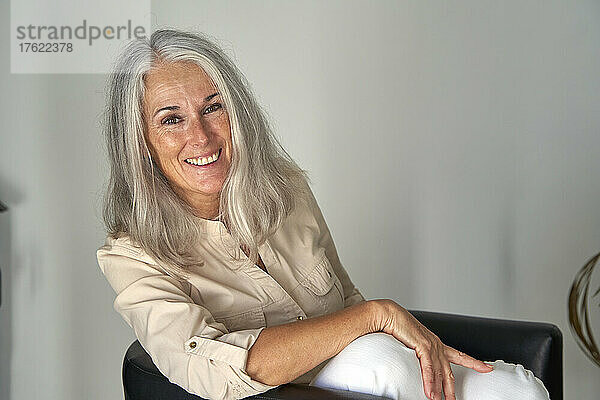 Glückliche Frau mit grauen Haaren sitzt auf einem Stuhl vor einer weißen Wand