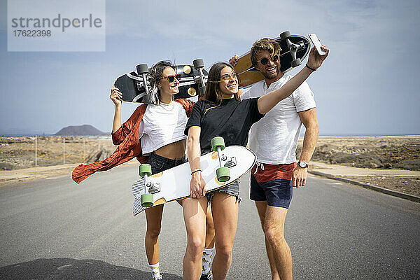 Glückliche junge Frau  die an einem sonnigen Tag ein Selfie mit Freunden macht  die Skateboards über ihr Mobiltelefon halten