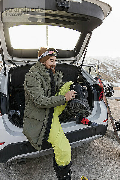 Lächelnder junger Mann im Kofferraum eines Autos  der sich auf das Snowboarden vorbereitet