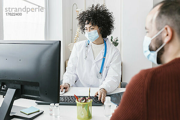 Arzt trägt Schutzmaske und sitzt am Computer mit Patient in der Klinik