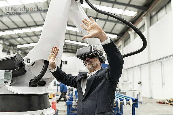 Geschäftsmann trägt Virtual-Reality-Simulator und steht mit erhobenen Armen in der Fabrik