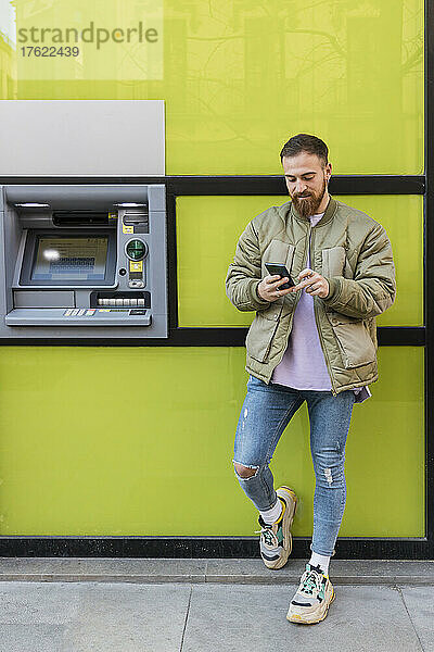 Junger Mann benutzt Smartphone und steht am Geldautomaten