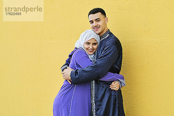 Lächelndes junges Paar  das sich umarmt und vor der gelben Wand steht