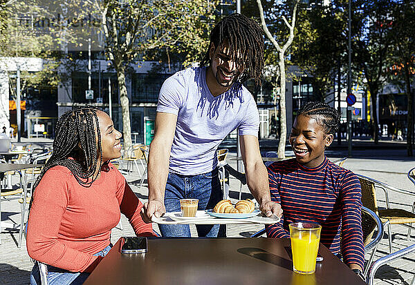 Glücklicher junger Mann trägt Tablett auf dem Tisch für Freunde im Straßencafé