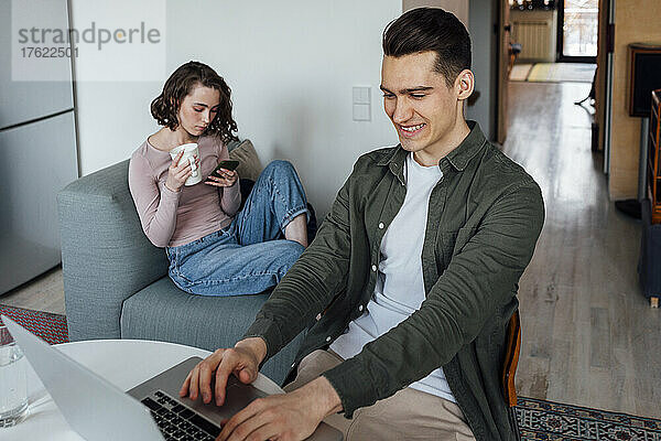 Lächelnder junger Mann mit Laptop und Frau  die zu Hause mit Kaffeetasse und Smartphone auf dem Sofa sitzt