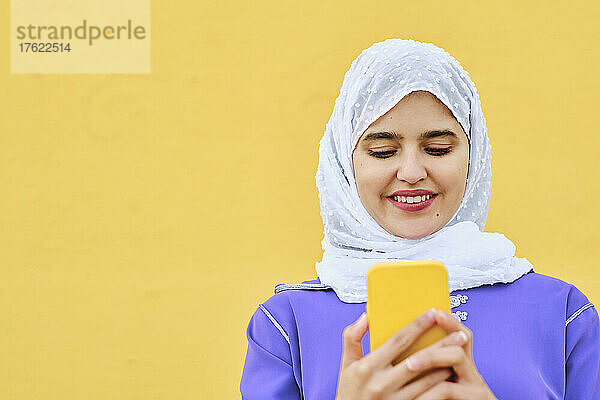 Smiling young woman surfing net through smart phone in front of yellow wall