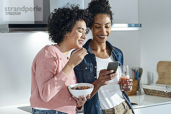 Lächelnde Frauen teilen ihr Smartphone in der heimischen Küche