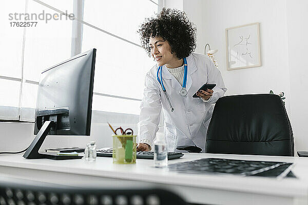Arzt hält Smartphone in der Hand und arbeitet am Computer in der Klinik