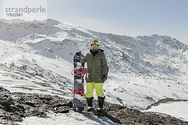 Lächelnder junger Mann steht mit Snowboard auf Felsen vor schneebedecktem Berg