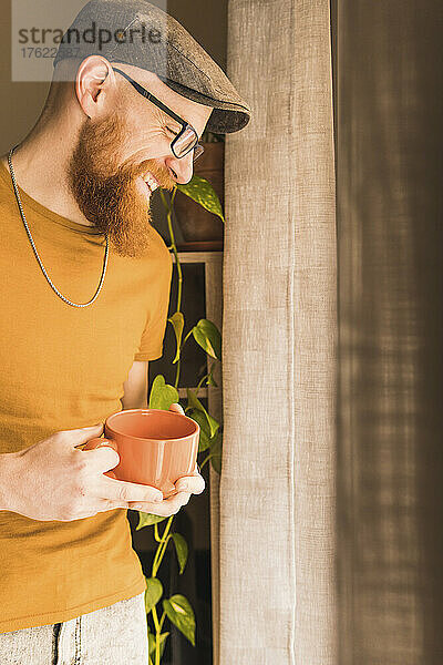 Cheerful man holding coffee cup standing by window at home