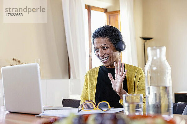 Lächelnde Geschäftsfrau mit Kopfhörern winkt bei Videoanruf über Laptop zu Hause