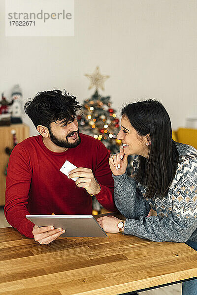 Lächelnder Mann hält Tablet-PC und Kreditkarte in der Hand und blickt seine Freundin zu Hause mit der Hand am Kinn an