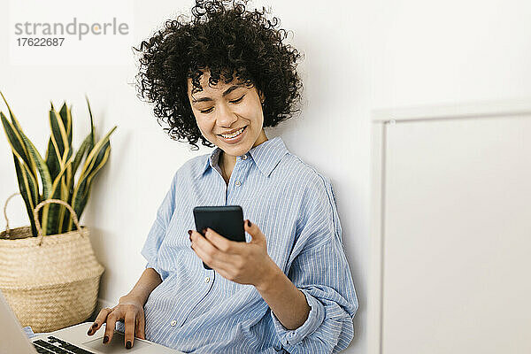 Lächelnde junge Frau mit Laptop und Smartphone sitzt zu Hause