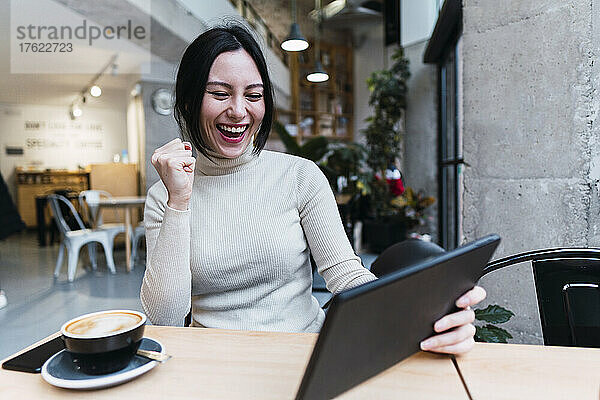 Fröhliche Frau gestikuliert mit der Faust und hält einen Tablet-PC im Café