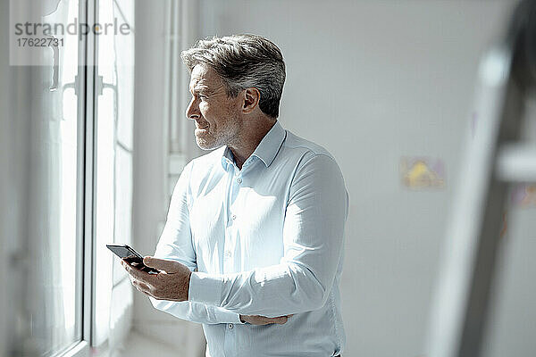 Geschäftsmann mit Smartphone blickt durch Fenster im Büro