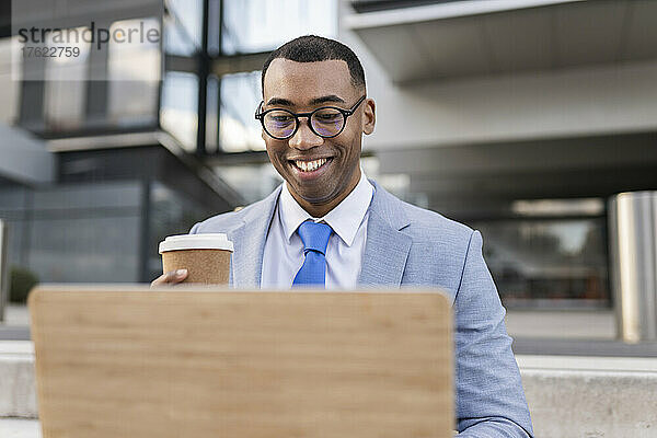 Glücklicher Geschäftsmann mit Einwegkaffeetasse und Laptop vor dem Gebäude sitzend