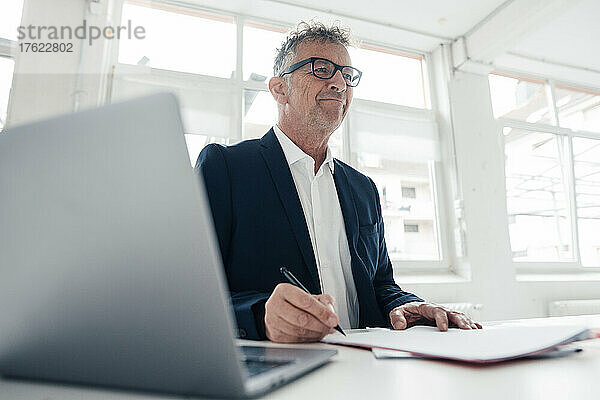 Lächelnder Geschäftsmann mit Laptop und Dokumenten auf dem Schreibtisch im Büro