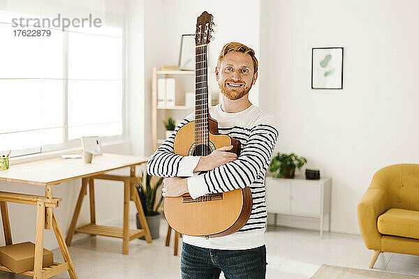 Glücklicher junger Mann mit Gitarre steht zu Hause im Wohnzimmer